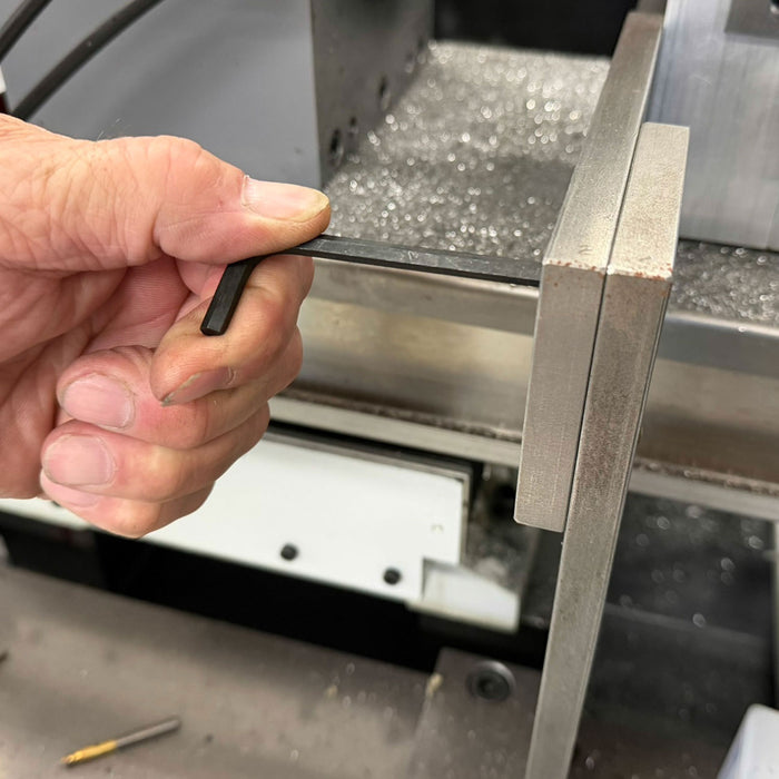 Hand holding a small black hex key near a metal surface with a machine in the background