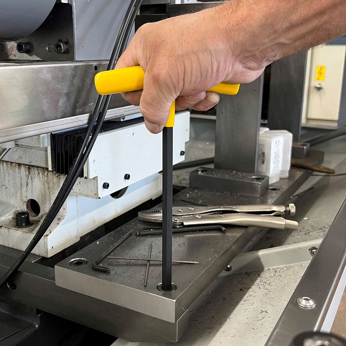 Hand using a yellow-handled tool on a metal workbench with equipment in the background