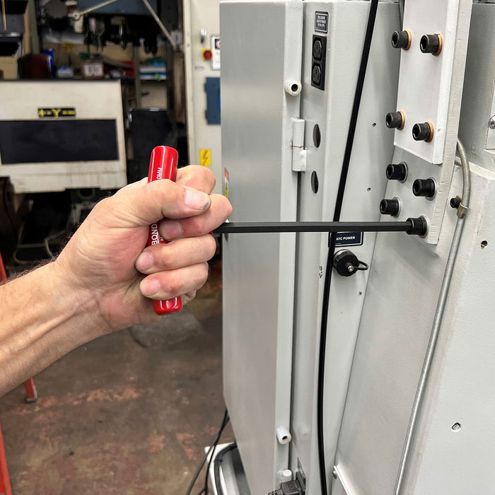 Hand holding a red handled tool near a large industrial machine in a workshop setting