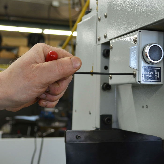 Hand holding a red handled tool near a release tool button on machine in an industrial setting